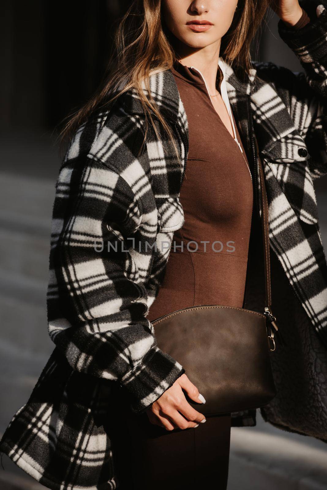 Outdoor fashion portrait of young beautiful fashionable girl wearing trendy checkered shirt, solid long sleeve bodycon one piece jumpsuits and small brown color small bag, posing in street.