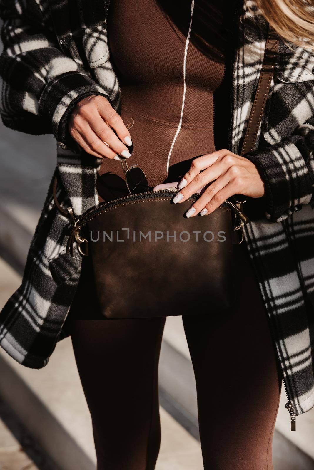 Outdoor fashion portrait of young fashionable girl wearing trendy checkered shirt, solid long sleeve bodycon one piece jumpsuits holding small brown color small bag.