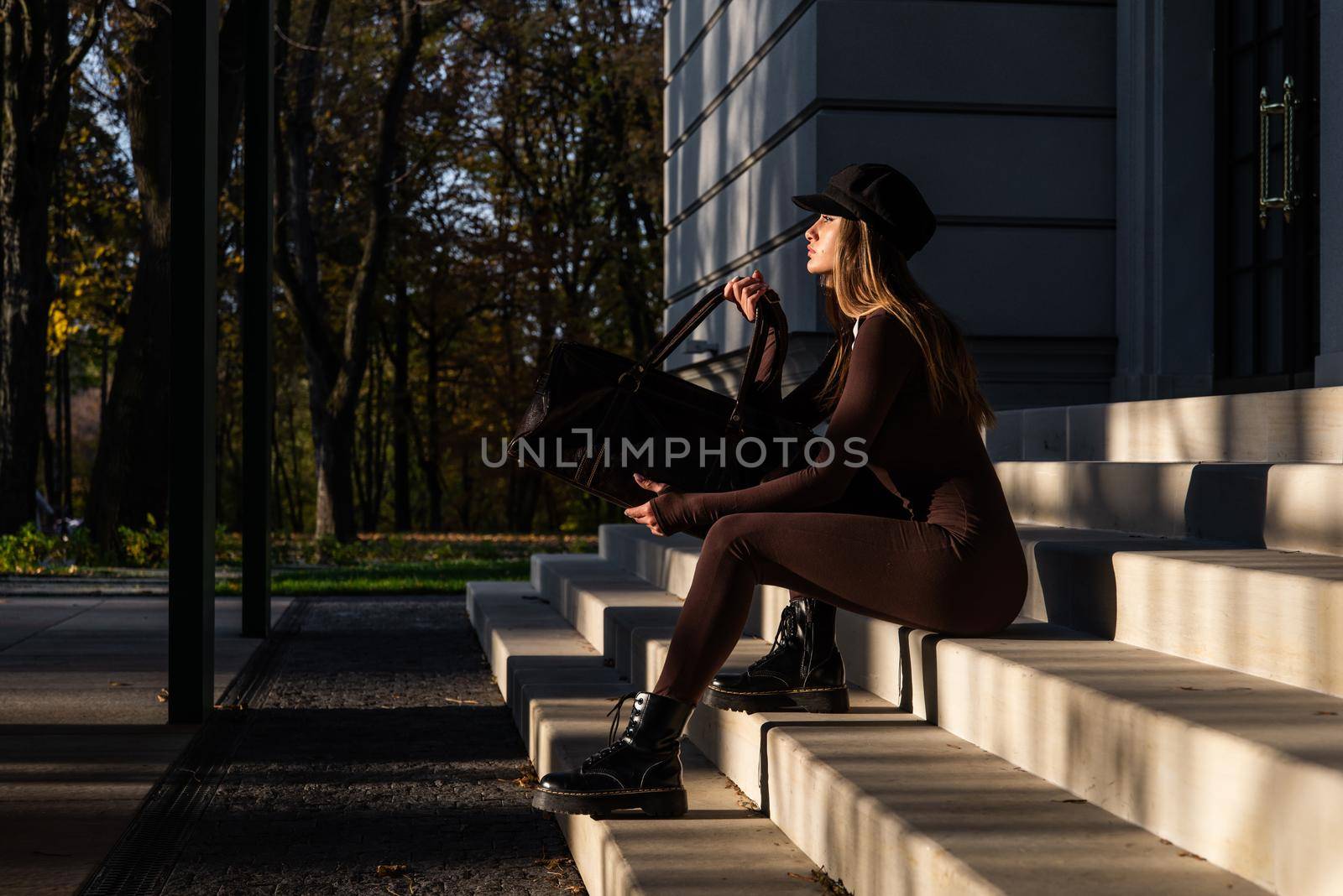 Fashionable young brunette woman with long legs. wearing solid long sleeve bodycon one piece jumpsuits posing with a leather brown travel bag on city street in old town