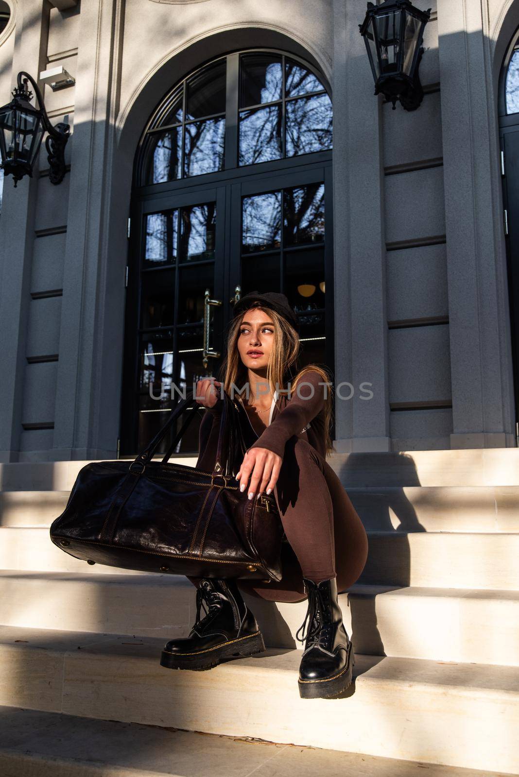 Fashionable young brunette woman with long legs wearing solid long sleeve bodycon one piece jumpsuits posing with a leather brown travel bag by Ashtray25