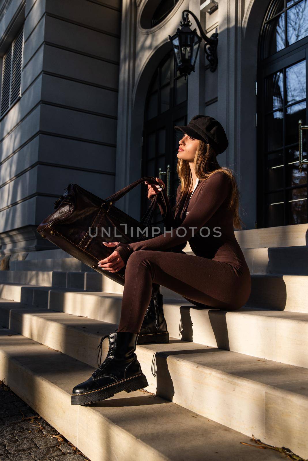 Fashionable young brunette woman with long legs. wearing solid long sleeve bodycon one piece jumpsuits posing with a leather brown travel bag on city street in old town