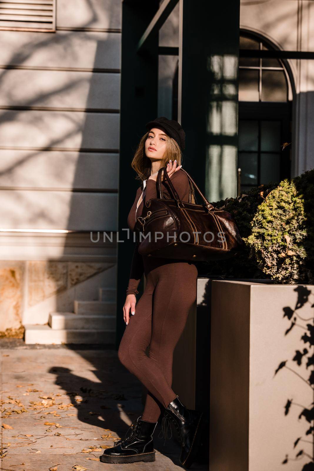 Fashionable young brunette woman with long legs. wearing Solid Long Sleeve Bodycon One Piece Jumpsuits posing with a leather brown travel bag on city street in old town