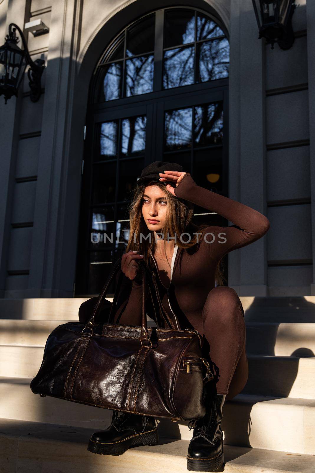 Fashionable young brunette woman with long legs. wearing solid long sleeve bodycon one piece jumpsuits posing with a leather brown travel bag on city street in old town