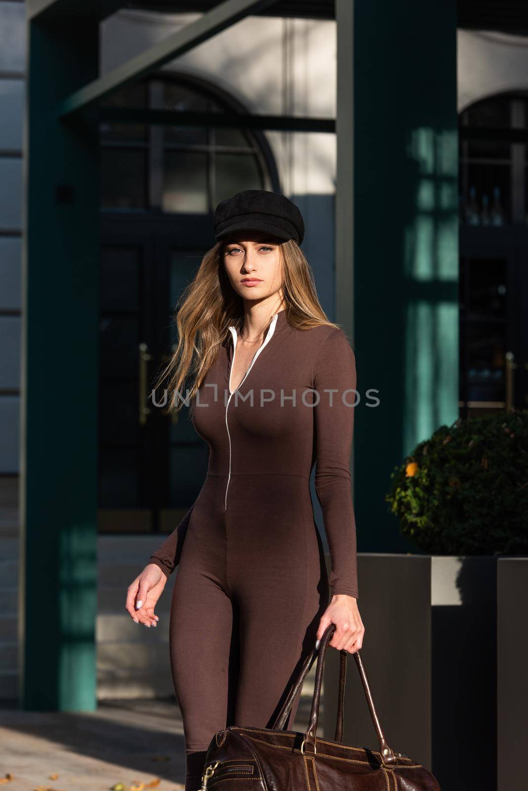 Fashionable young brunette woman with long legs. wearing Solid Long Sleeve Bodycon One Piece Jumpsuits posing with a leather brown travel bag on city street in old town