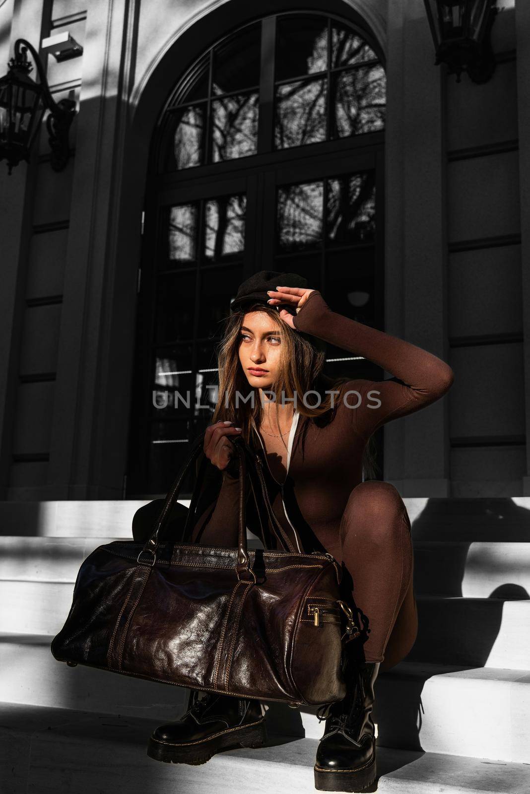 Fashionable young brunette woman with long legs. wearing solid long sleeve bodycon one piece jumpsuits posing with a leather brown travel bag on city street in old town