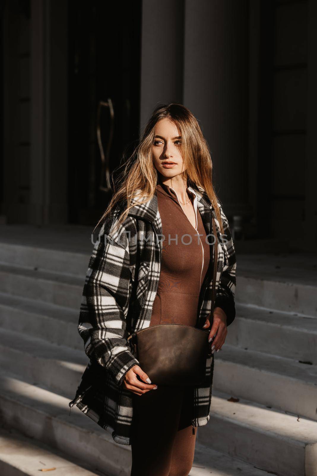 Outdoor fashion portrait of young beautiful fashionable girl wearing trendy checkered shirt, solid long sleeve bodycon one piece jumpsuits and small brown color small bag, posing in street.