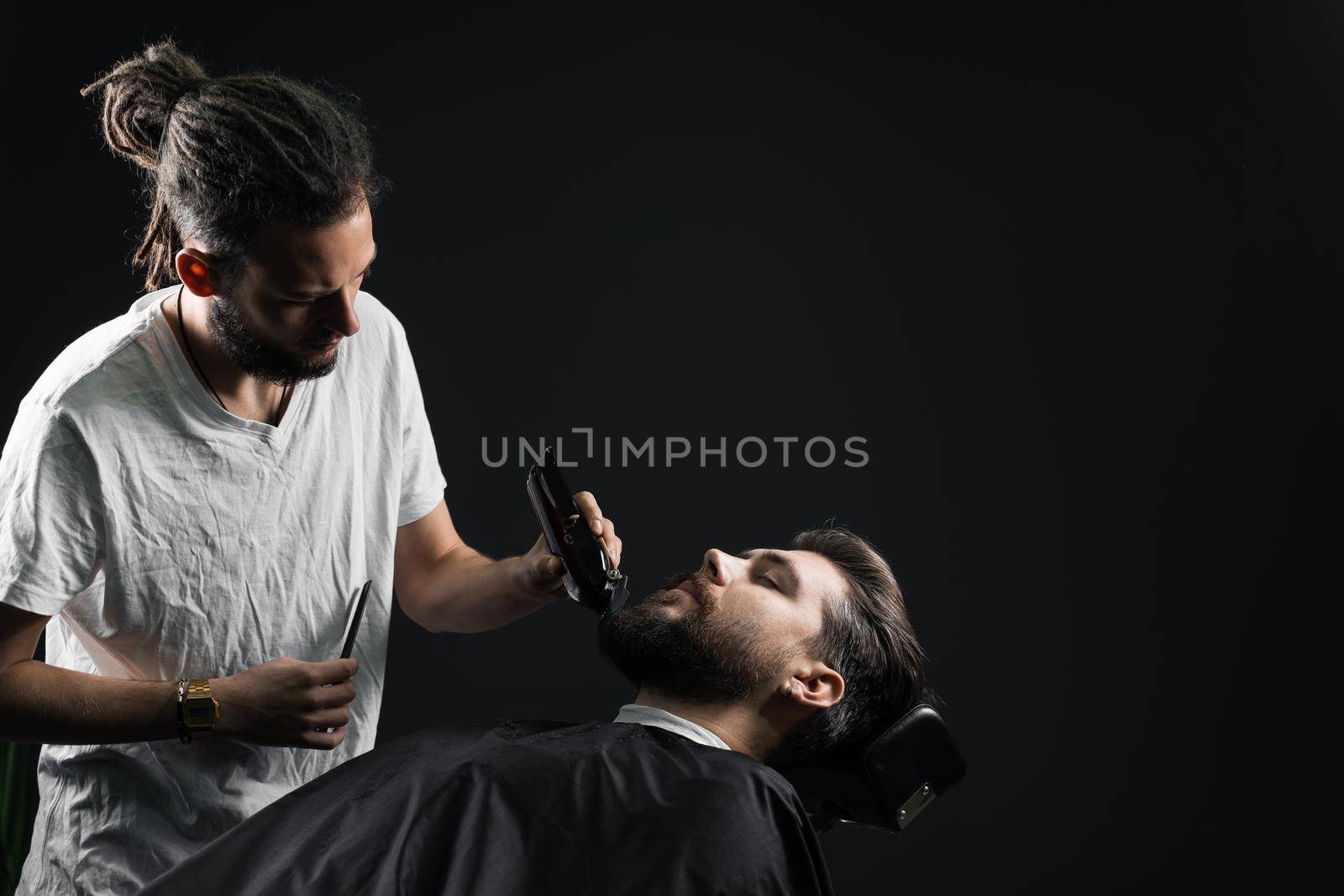 Beard shaving in barbershop. Barber with dreadlocks trim handsome bearded man