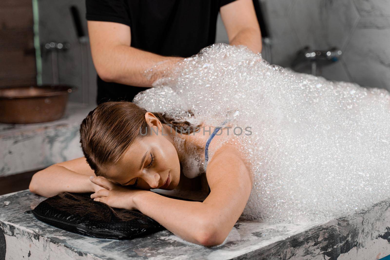 Foam peeling in Turkish spa hammam. Attractive model is relaxing in spa. Masseur is making massage using sponge glove. by Rabizo