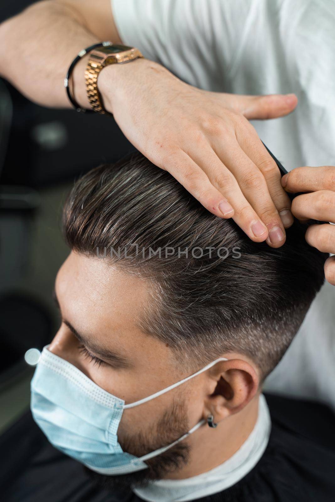 Hair styling with wax for a bearded man in a medical mask. The work of the barbershop in quarantine coronavirus covid-19.