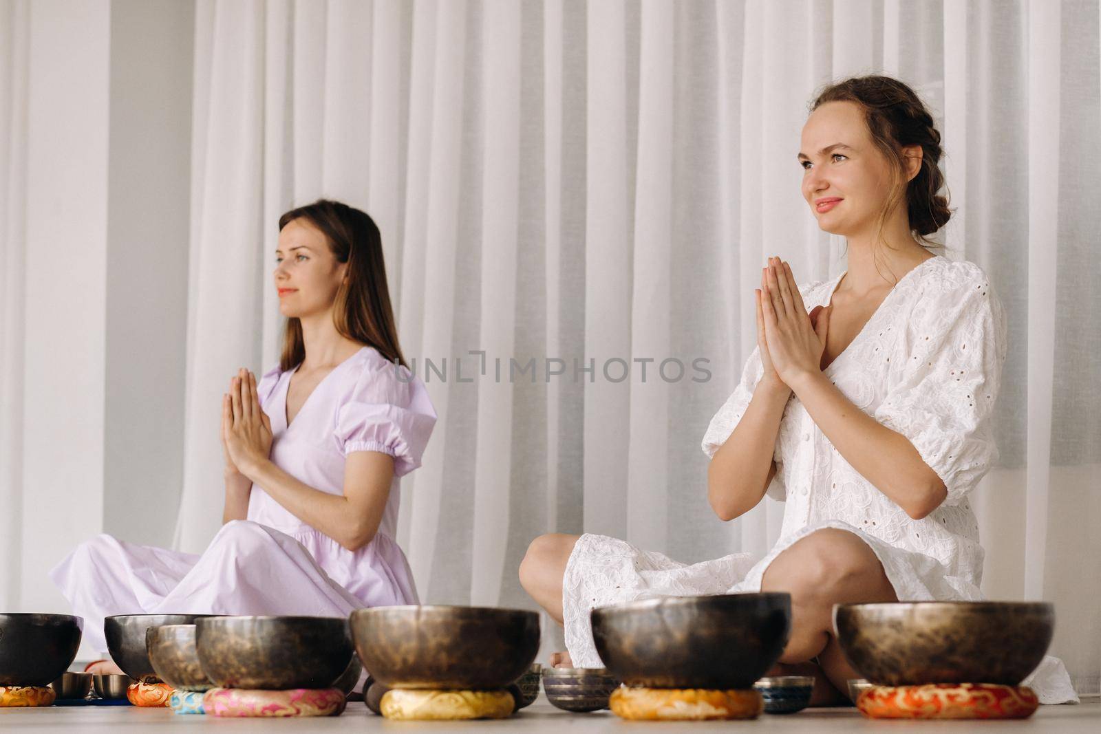Two women are sitting with Tibetan bowls in the lotus position before a yoga class in the gym by Lobachad