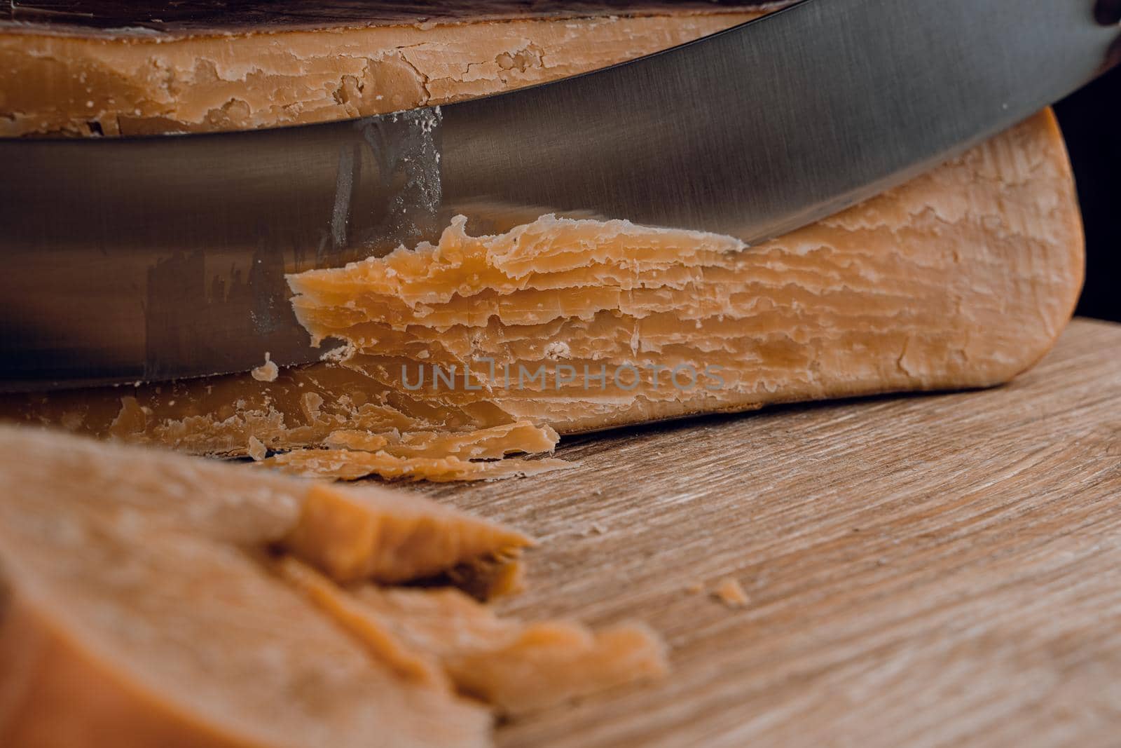 Slicing aged cheese parmesan with crystals using a cheesy dutch knife. Hard cheese with knife on dark background. by Rabizo