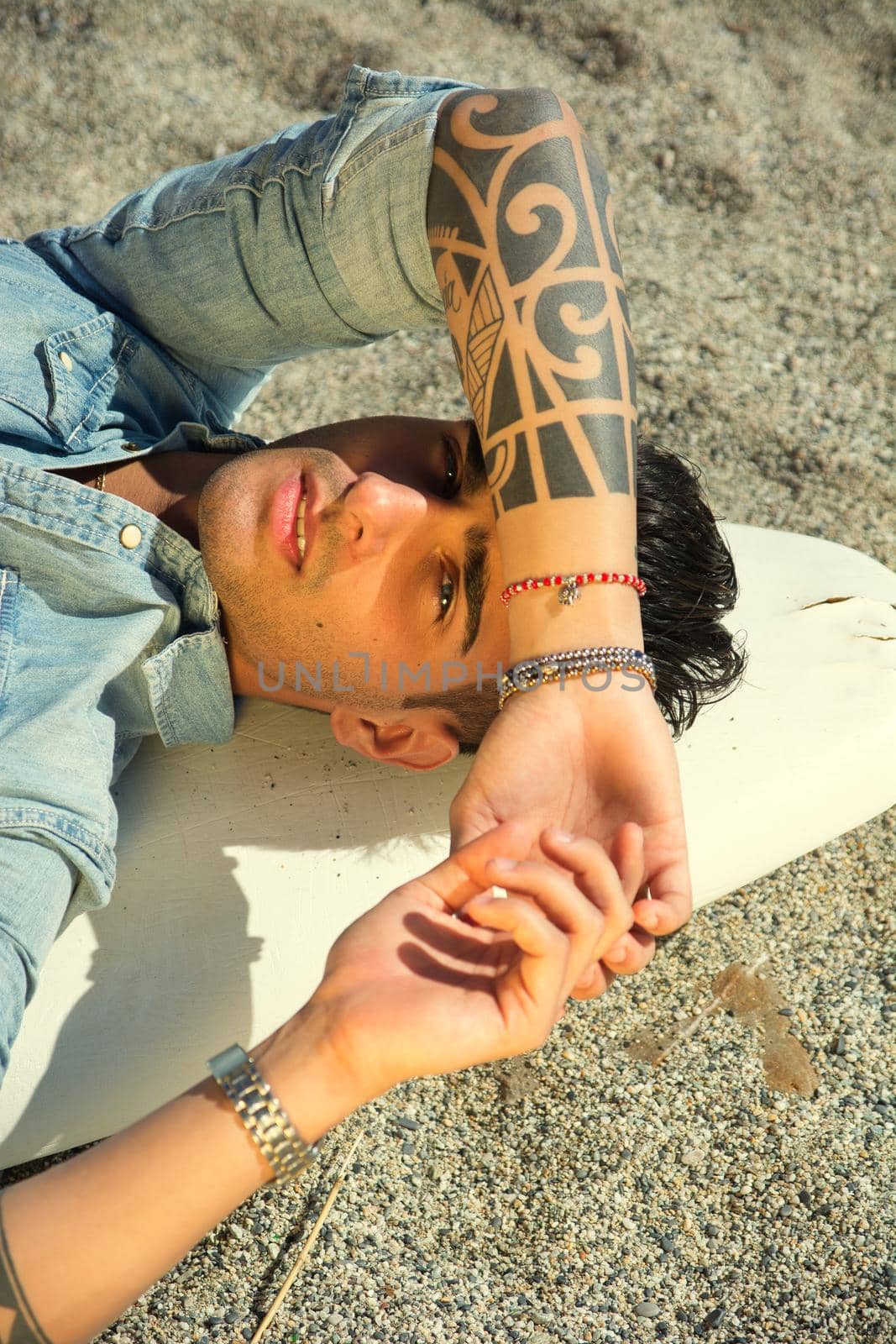 Stylish handsome young man laying on surfboard on beach in sunlight