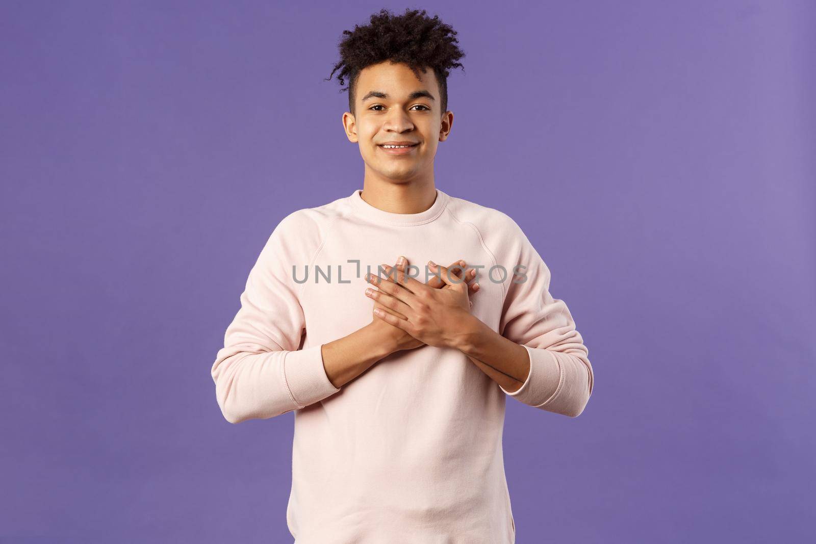 Portrait of romantic handsome hispanic man with dreads, hold hands pressed to heart and smiling pleased, see heartwarming nice gesture, receive lovely gift, being touched, purple background.