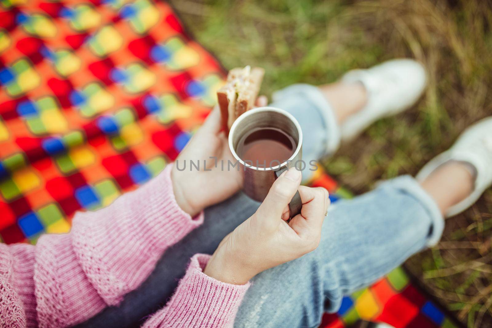 Snack in nature outdoors. Close up shot of female hand holding sandwich and cup of hot drink. Caucasian woman sitting on picnic blanket at grass. Top view.