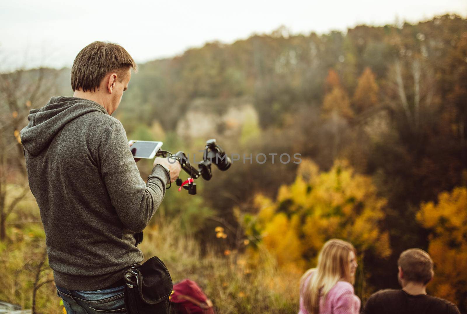 Young man going to shoot couple of tourists in love sitting on top of mountain in Autumn nature. Rear view of cameraman preparing camera on digital stabilizer for shooting photo or video.