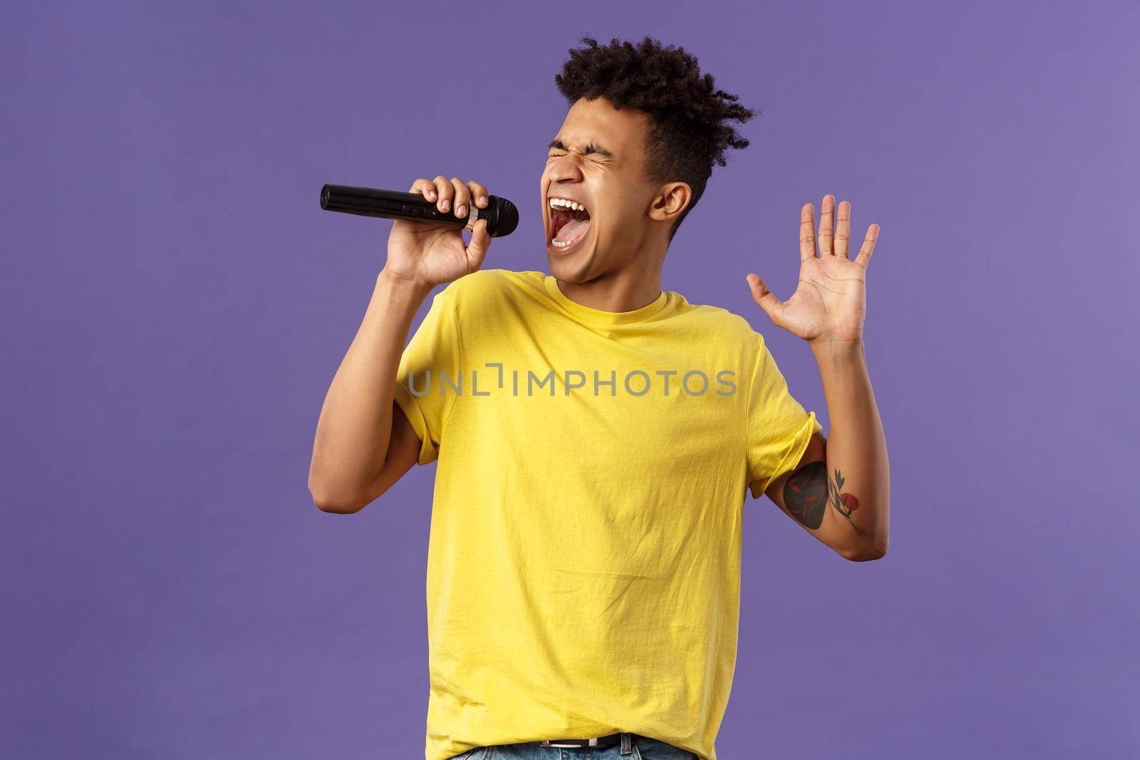 Portrait of passionate carefree young hispanic singer with dreads and tattoos, reaching highest note in song, raising hand up singing loud at microphone with closed eyes, purple background.