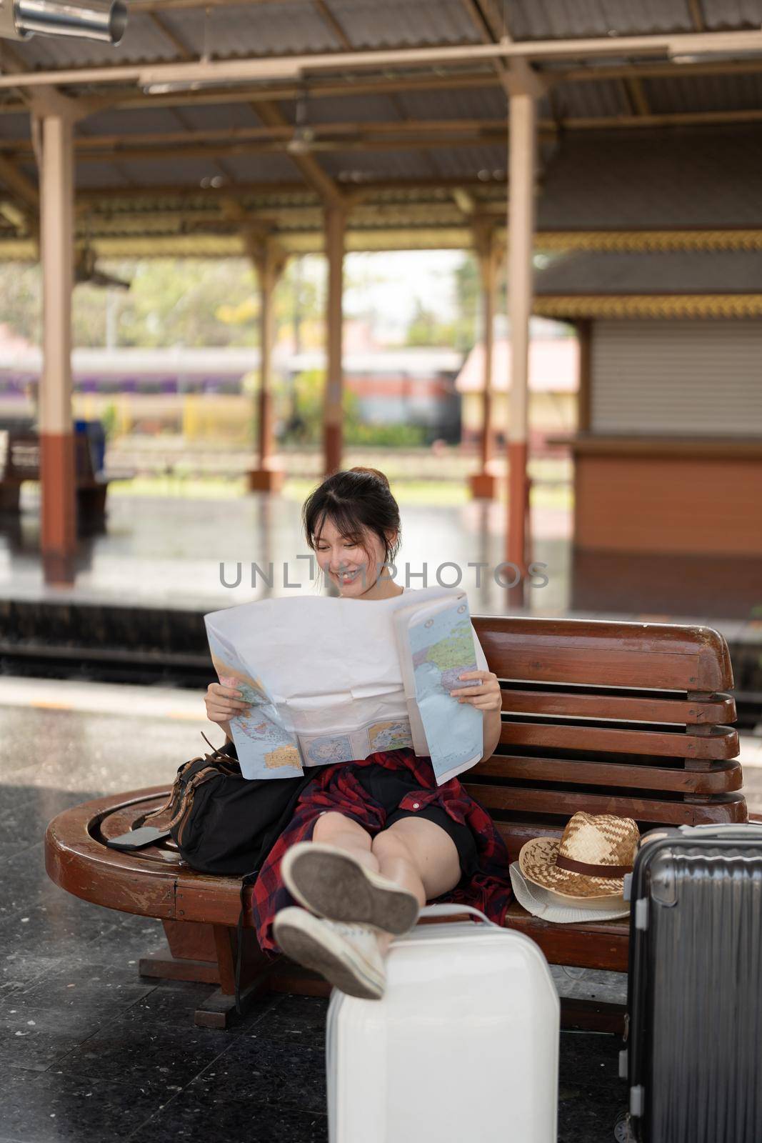 Happy young asian woman traveler or backpacker using map choose where to travel with luggage at train station, summer vacation travel concept by nateemee