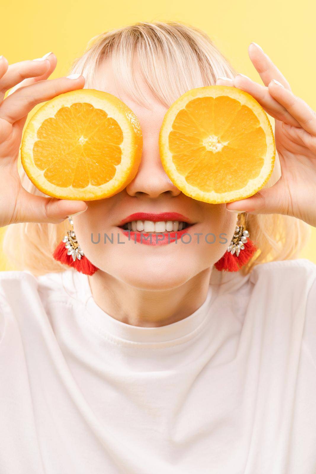 A woman with oranges in her eyes. Red lipstick lips. Merry, cheerful girl exudes positive, spoiled oranges. Isolated on yellow background by StudioLucky