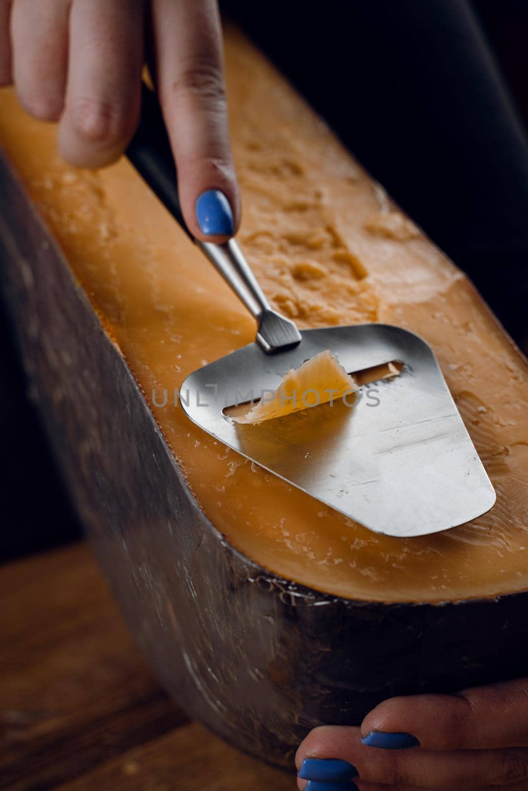 Slicing aged cheese parmesan with crystals using slicer knife. Hard cheese with knife for appetizer on dark background