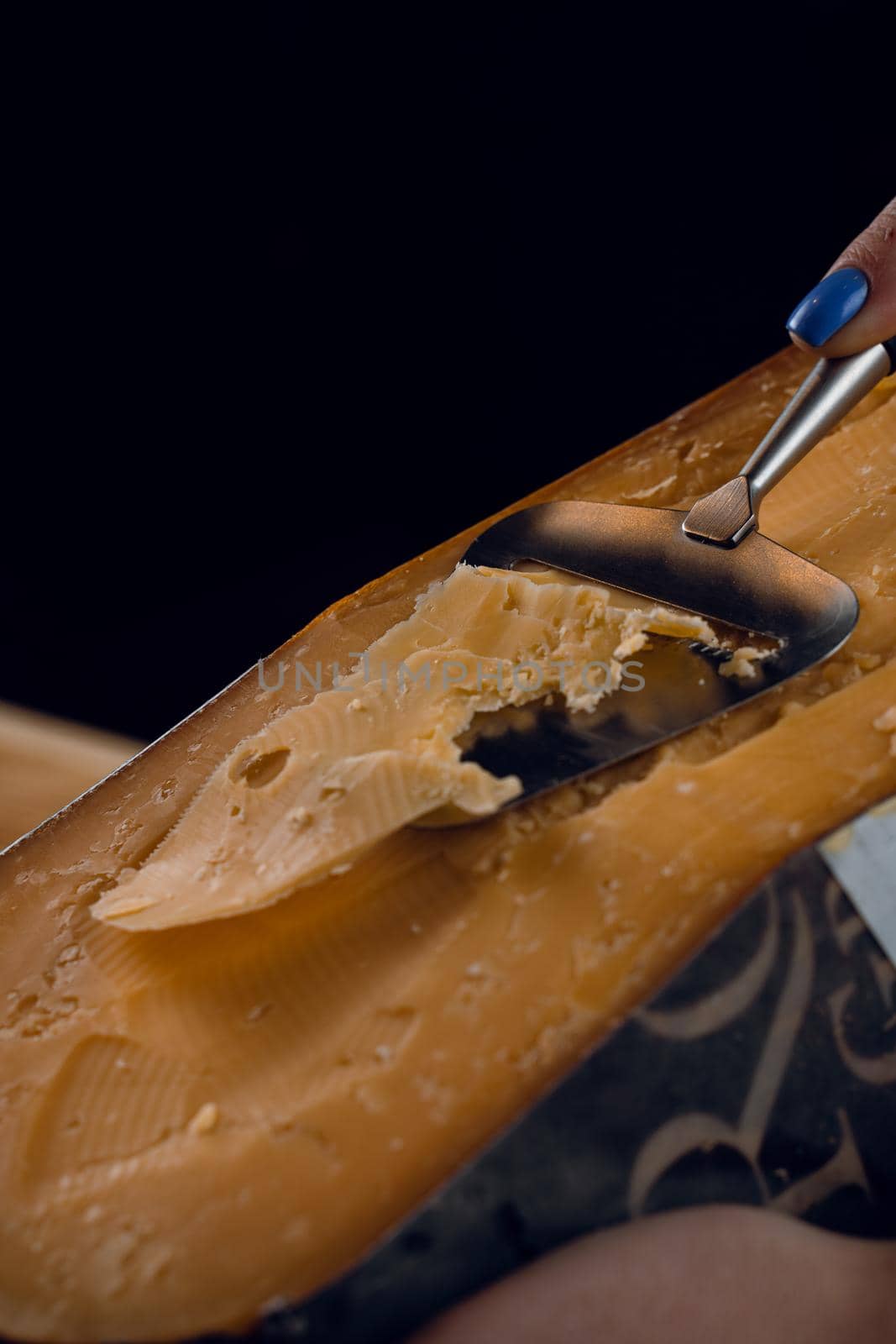 Slicing aged cheese parmesan with crystals using slicer knife. Hard cheese with knife for appetizer on dark background