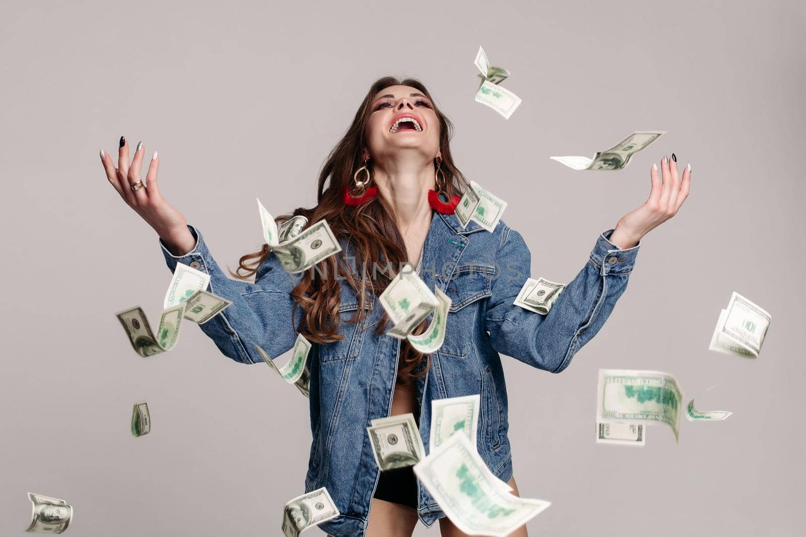 Cheerful and positive brunette woman with bright stylish earrings and denim jacket laughing with arms in the air in dollar banknotes flying around. Isolate on grey or white background. Studio shot.