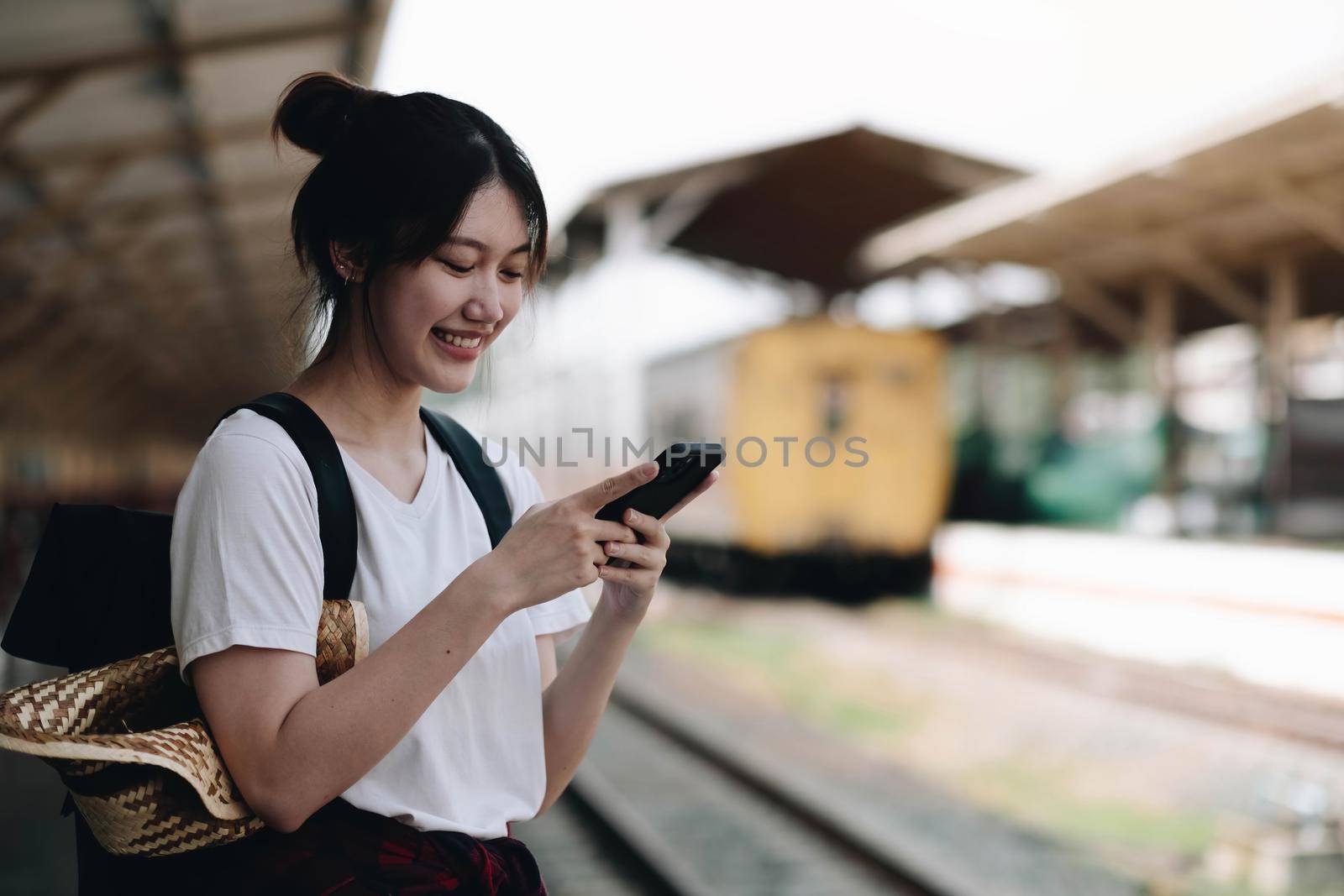 Woman waiting on station platform on background light train using smart phone. Tourist texting message and plan route of stop railway, railroad transport, booked. Enjoying travel concept by wichayada