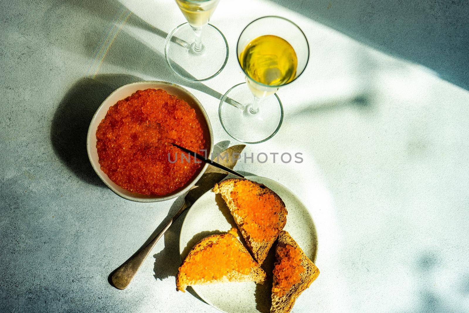 Red trout caviar in a bowl and sparkling wine in glasses on concrete background with deep shadows in sunny day