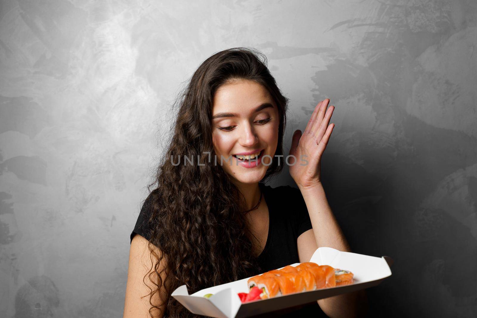 Surprised girl with sushi set philadelphia rolls in a paper box happy girl holding on a gray background. Food delivery