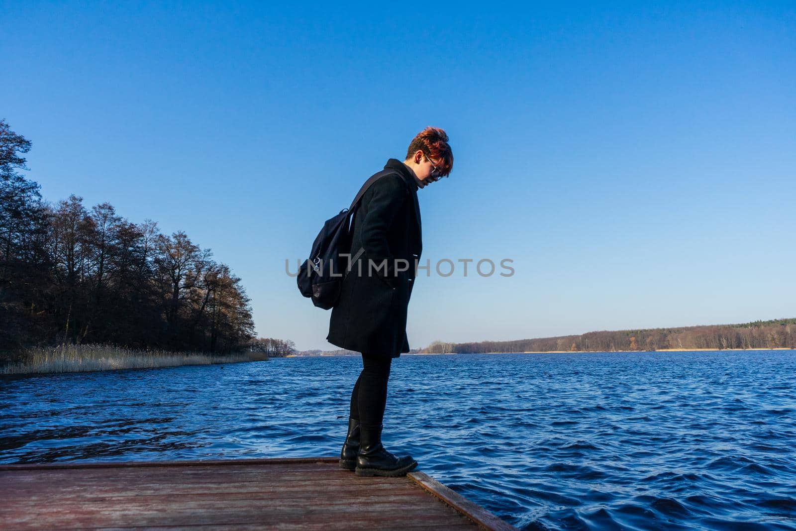 girl in a black coat stands at the edge of the pier and looks into the water of the lake.