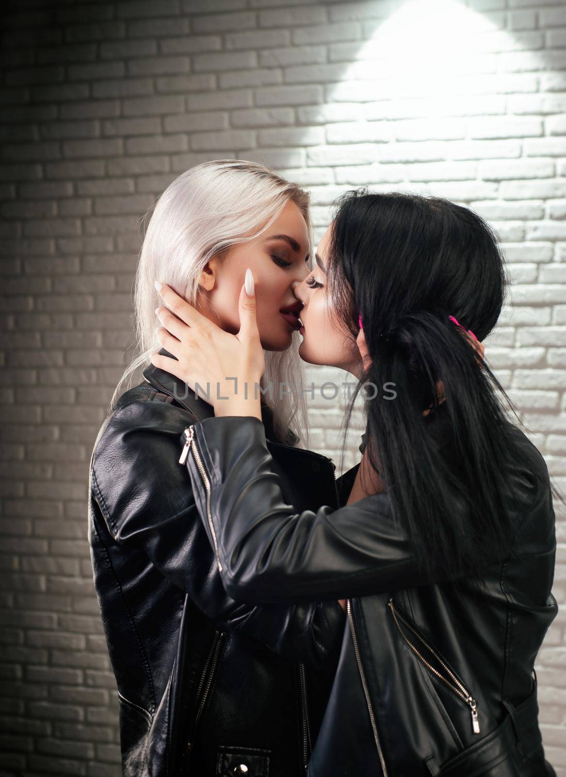 the two young lesbian lgbt women in leather jackets kiss on the lips against a white brick wall