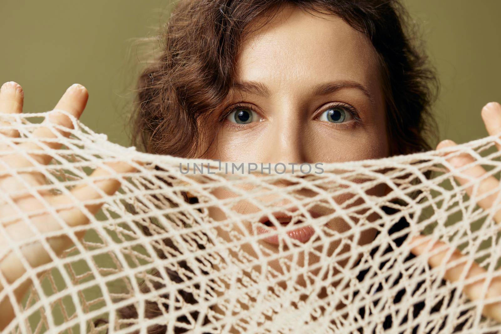 Conceptual Abstract Art. Excited curly beautiful female hold string bag near face seriously looks at camera posing isolated on olive green background. Eco-friendly Natural products concept. Copy space by SHOTPRIME