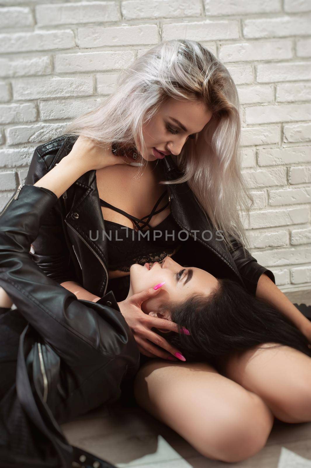 two young lesbians lustful women in leather jackets embrace against a white brick wall showing intimacy and flirtation by Rotozey