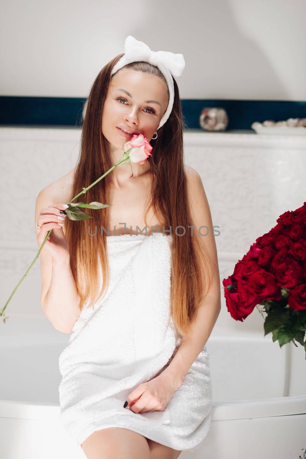 Brunette girl with long hair sitting on bath after morning shower. Pretty lady in white towel looking up and holding flower. Young woman getting bouquet of roses and thinking about boyfriend.