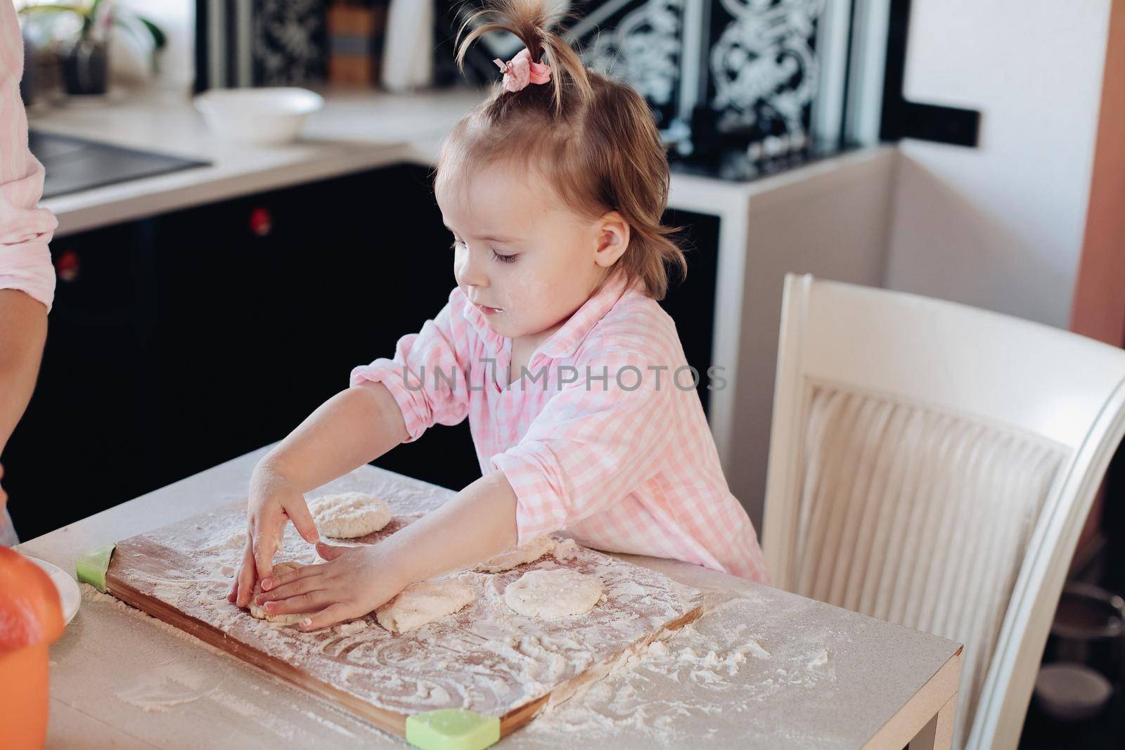 Close up of little serious kid sitting on white chair in pink pajama. Lovely daughter helping her mother preparing breakfast for family. Cute child in flour cooking together with parent at kitchen.