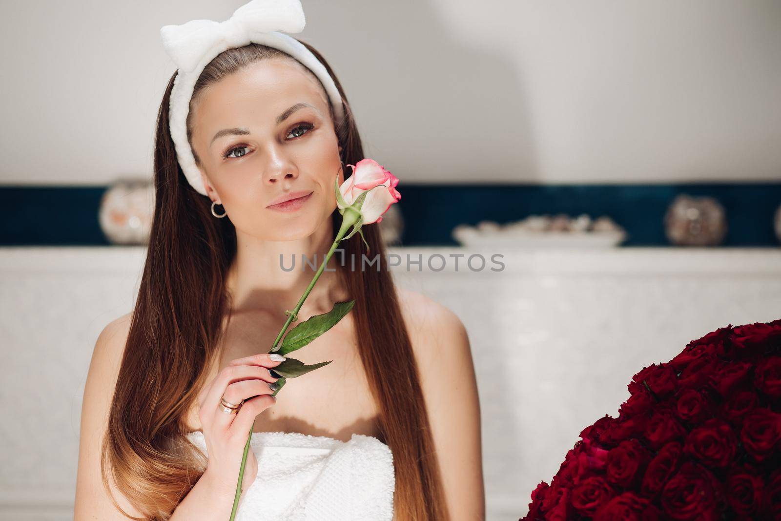 Portrait of sensual beautiful woman in white headband with bow for washing face smiling slightly at camera with fragile pink rose bud at her face.