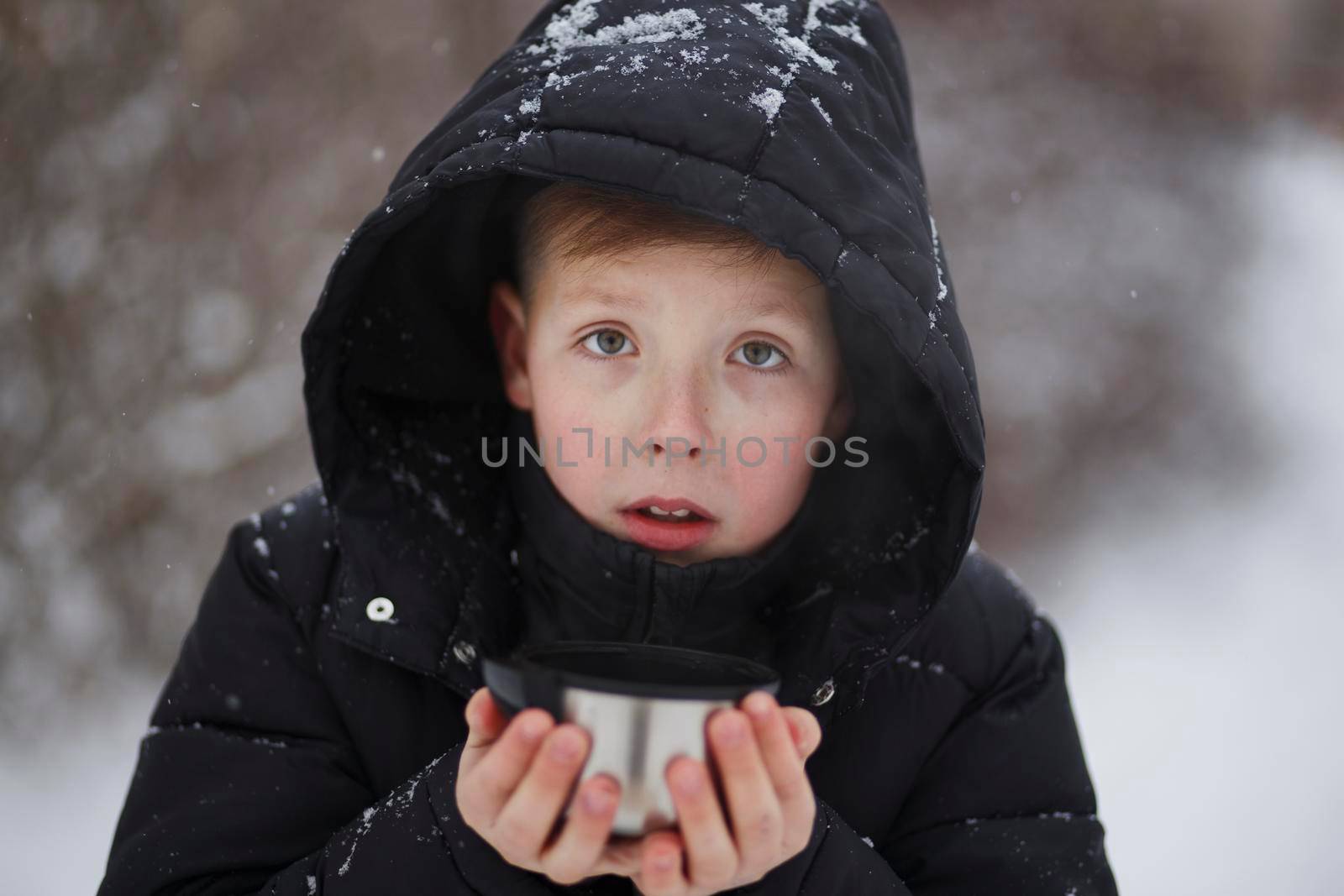 Portrait of a cute teenage boy in winter clothes with a hood holding a metal mug with tea in his hands. Warm yourself in nature in winter with hot tea.