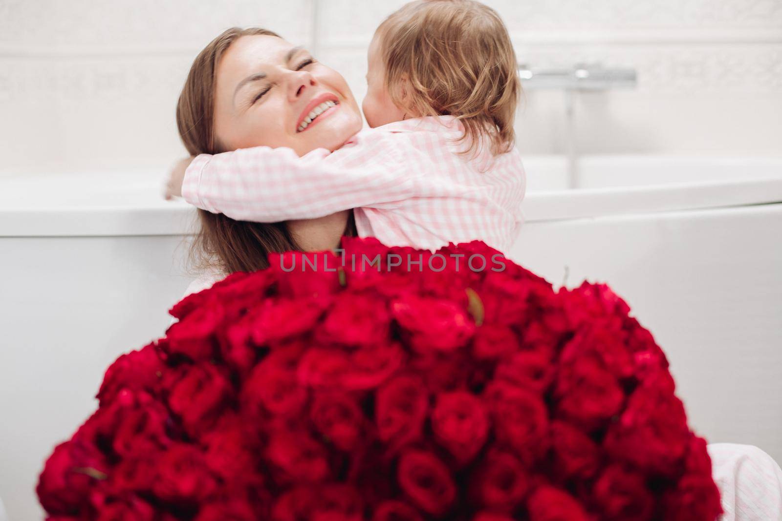 Beautiful young mother posing with little girl and big bouquet of red roses. Pretty woman kissing cute child and smiling. Happy family enjoying present. Concept of love and happiness.