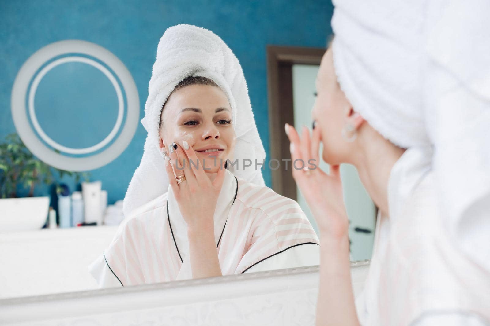 Beautiful woman in white coat and towel on head looking at mirror on her face. Young pretty lady standing in light bathroom after morning shower. Attractive girl using cream on skin before make up.