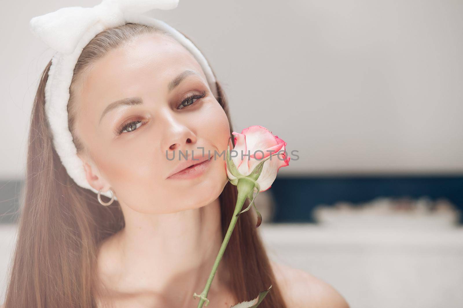 Portrait of sensual beautiful woman in white headband with bow for washing face smiling slightly at camera with fragile pink rose bud at her face.