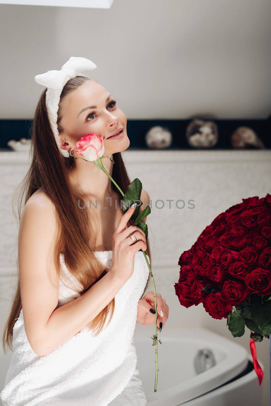 Brunette girl with long hair sitting on bath after morning shower. Pretty lady in white towel looking up and holding flower. Young woman getting bouquet of roses and thinking about boyfriend.