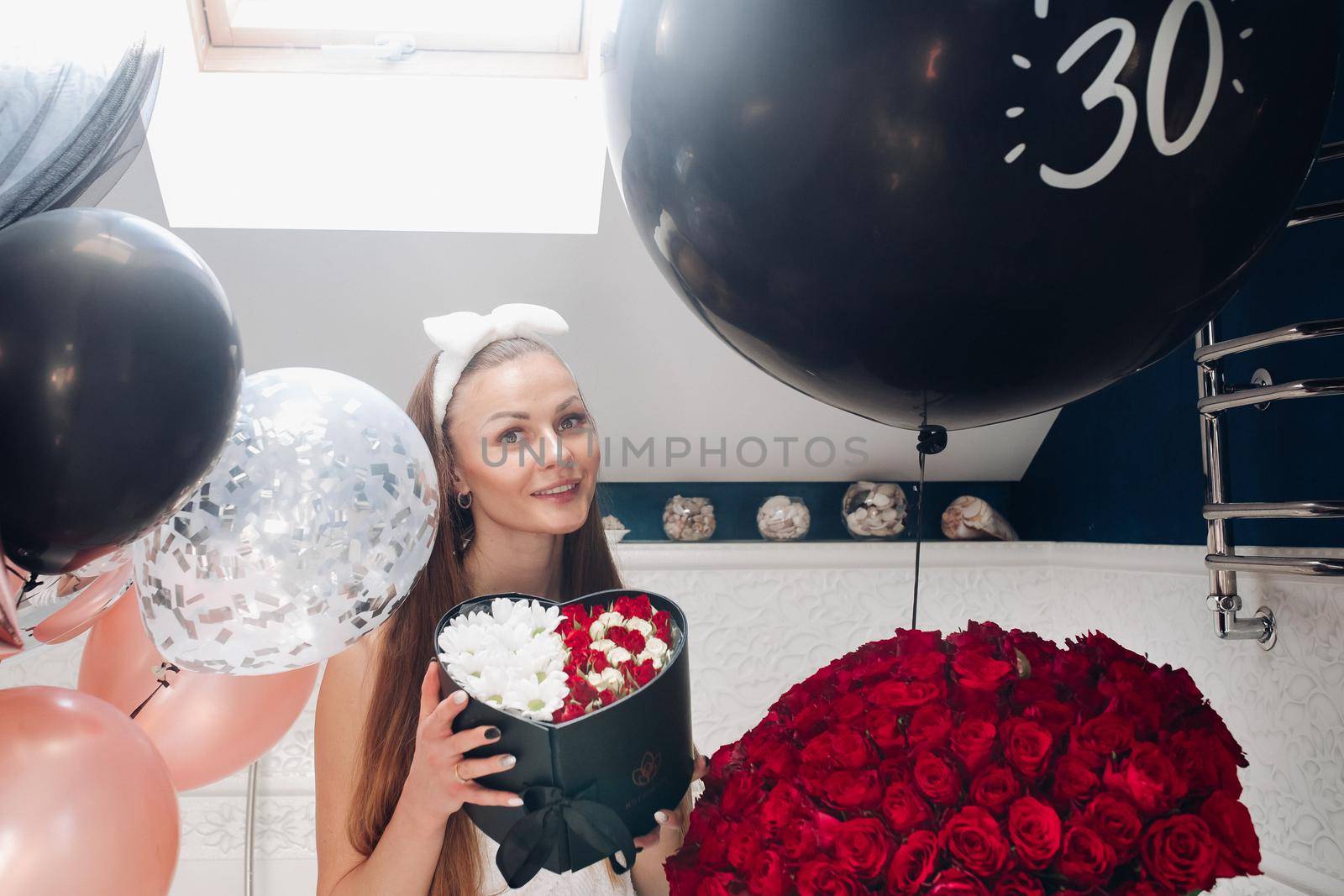 Portrait of positive beautiful brunette woman in headband and towel enjoying flowers in box surrounded by colorful air balloons and bouquet of red roses. She is in the bathroom recieving presents.