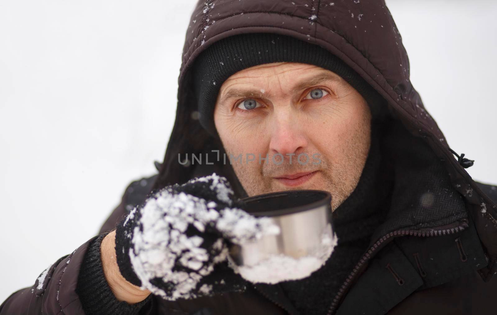 Portrait of a handsome unshaven man in winter clothes with a hoodie wearing black gloves holding a metal mug. Portrait of a man 40-55 years old with a mug against a background of snow.