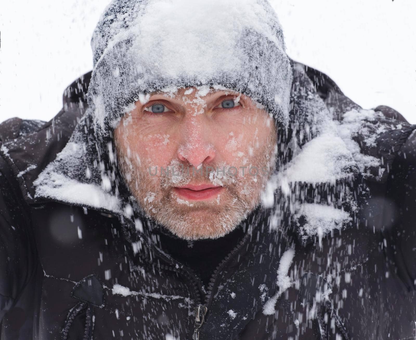 A man in a winter jacket and a black hat covered in snow. Portrait of a tourist in the snow during bad weather. Freezing man in the snowfall.