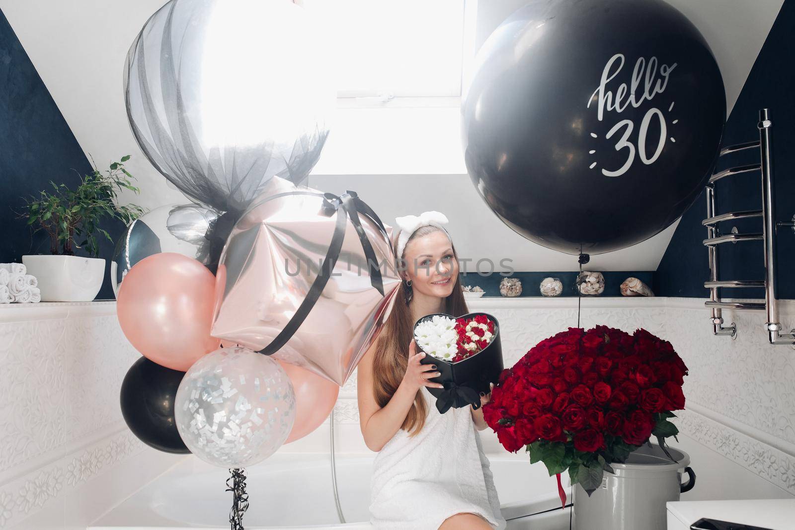 Portrait of positive beautiful brunette woman in headband and towel enjoying flowers in box surrounded by colorful air balloons and bouquet of red roses. She is in the bathroom recieving presents.