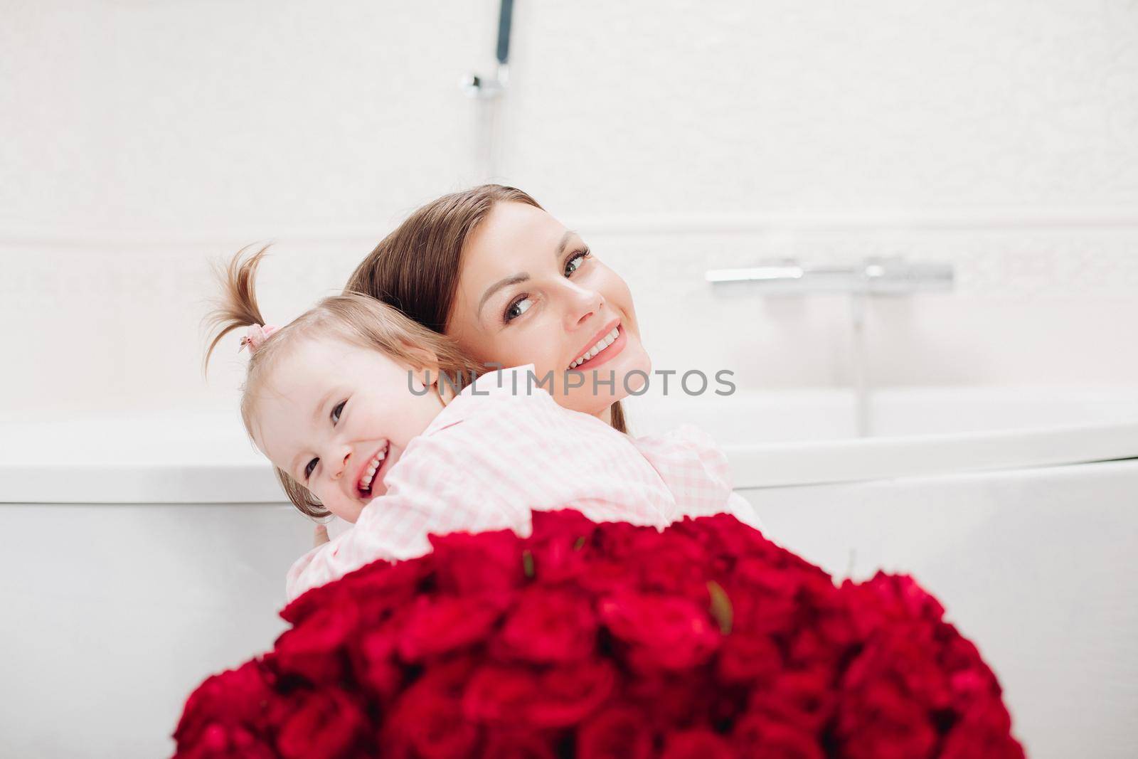 Happy mother and cute daughter sitting in bathroom in morning and smiling. Brunette woman with long hair holding lovely child and looking up. Young wife getting red roses from husband on holiday.