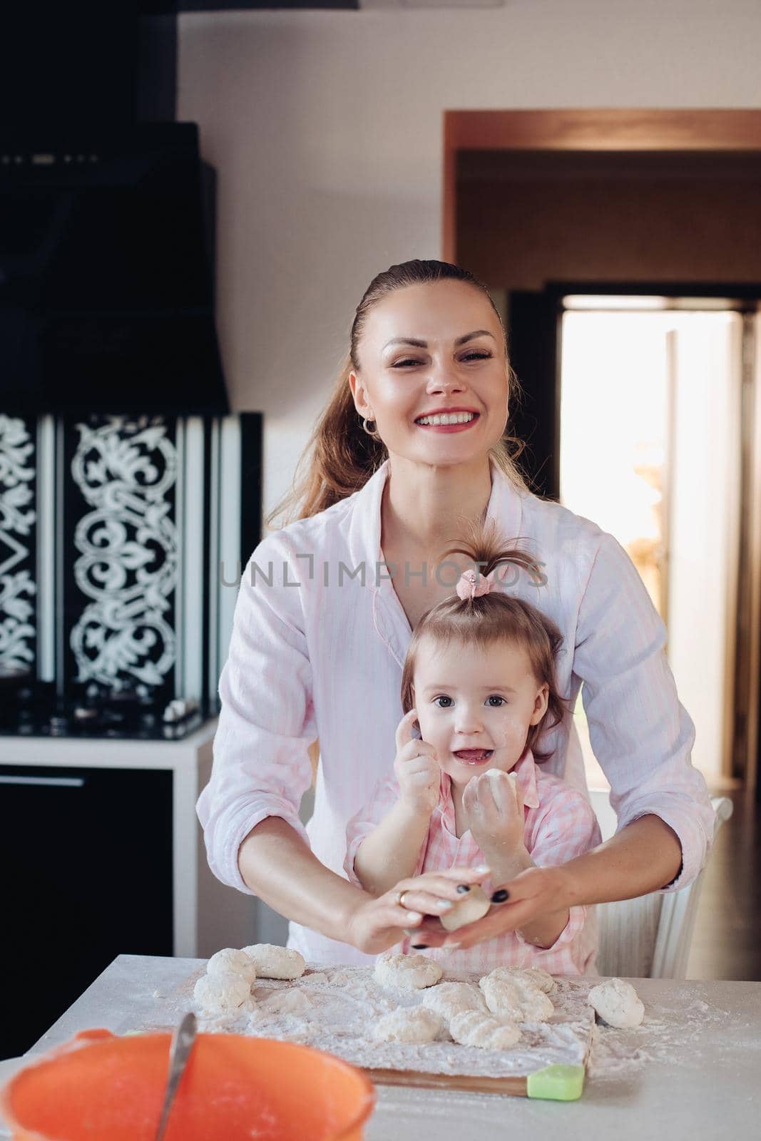 Portrait of gorgeous young adult mother in shirt cooking with her little daughter in similar pink plaid shirt. They are smiling while kneading dough in the kitchen.