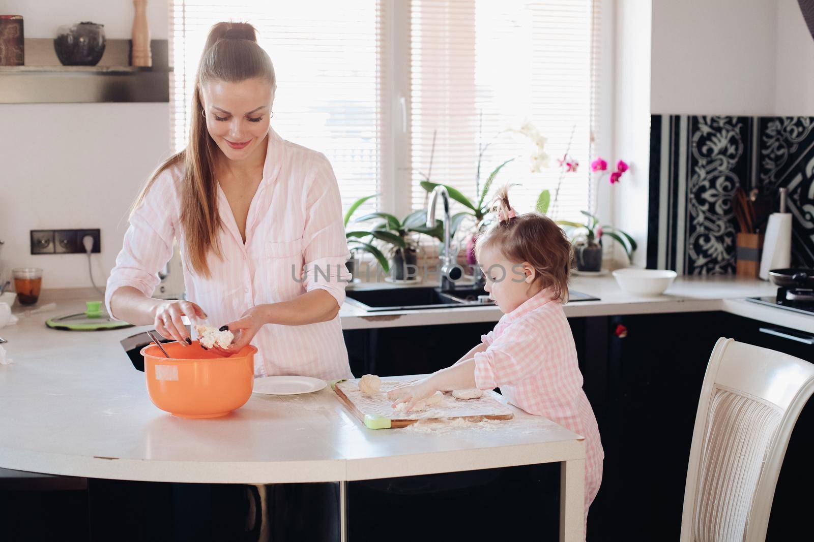 Happy wife smiling and preparing breakfast together with child for husband. Beautiful mother with cute daughter cooking in pajamas in morning. Little lovely kid helping parent with dough at kitchen.