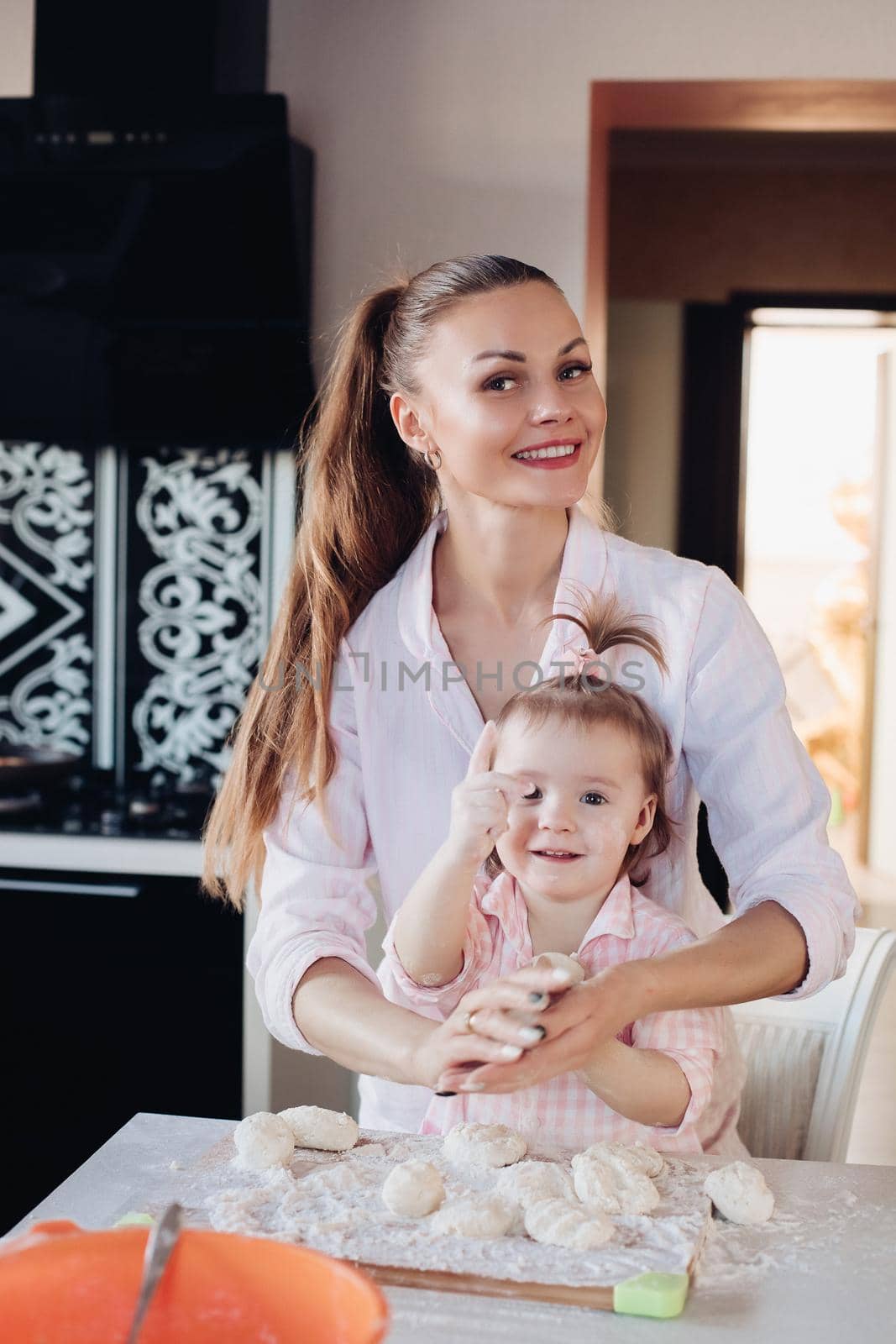 Portrait of gorgeous young adult mother in shirt cooking with her little daughter in similar pink plaid shirt. They are smiling while kneading dough in the kitchen.