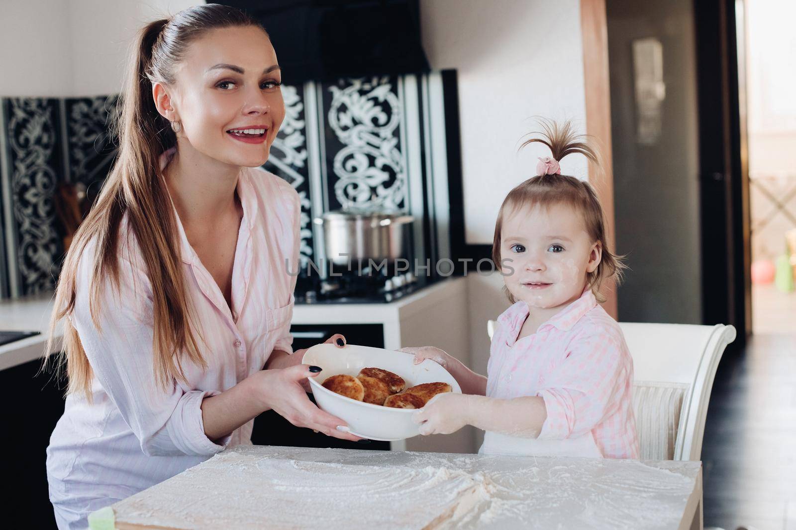 Beautiful woman with lovely child in pajamas holding big bowl with cookies. Close up of happy mother and daughter sitting together at kitchen in morning. Little kid helping parent cooking breakfast.