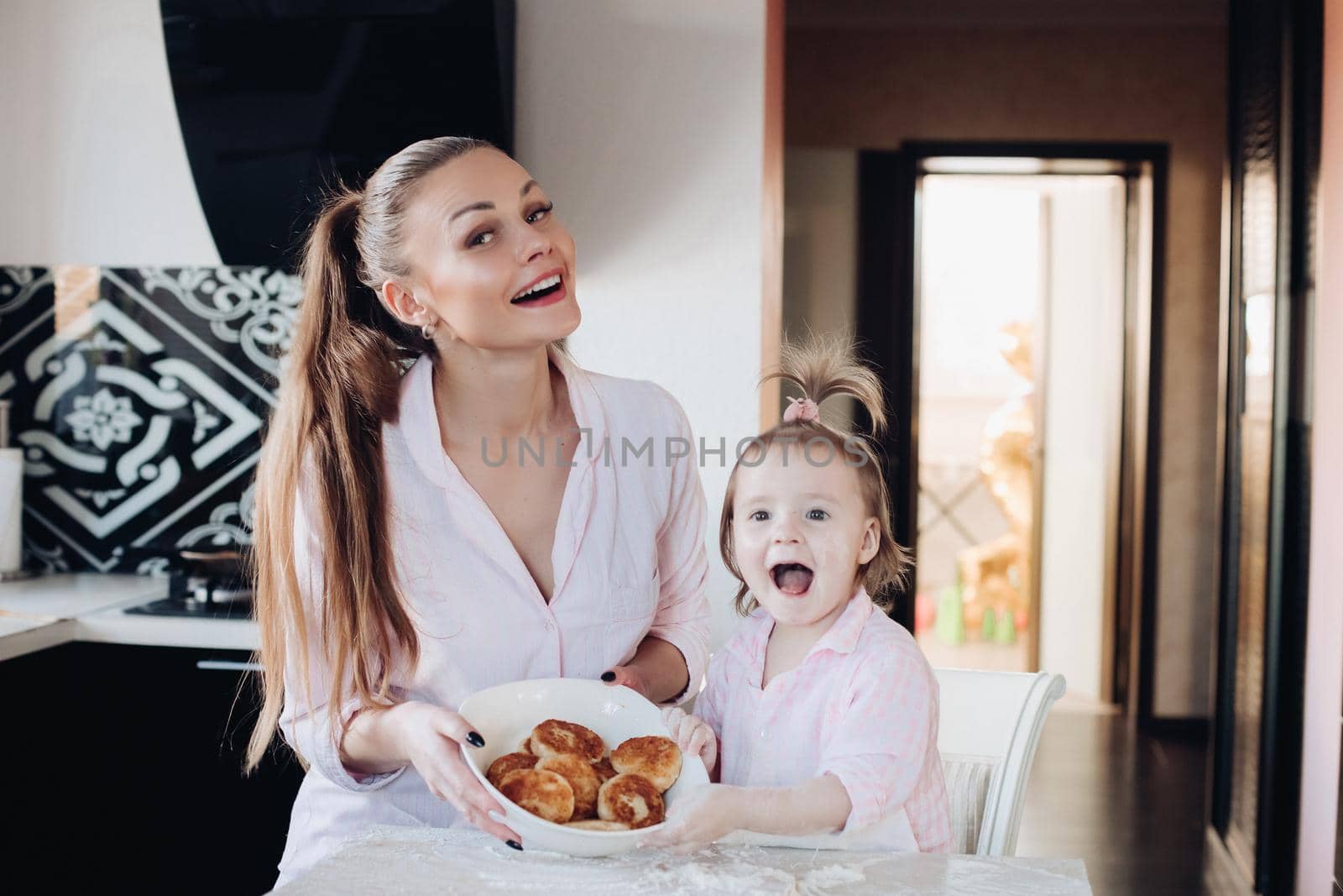 Front view of attractive mother and little daughter looking at camera and showing baked curd fritter. Happy family cooking and playing together in kitchen. Concept of love and happiness.